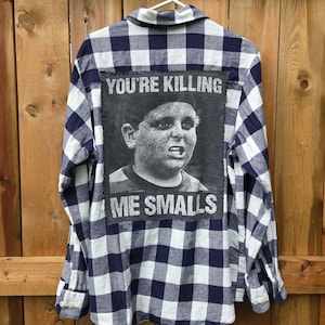 Puede incluir: Una camisa de manga larga con botones y un patrón de cuadros azul marino y blanco. La parte posterior presenta un parche de tela grande, rectangular y gris oscuro con un retrato en blanco y negro de un niño y el texto YOU'RE KILLING ME SMALLS.