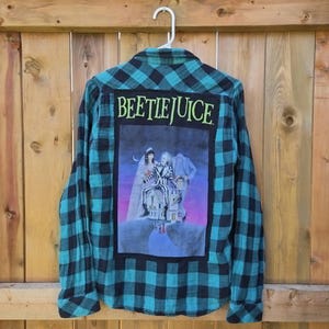 Może przedstawiać: Koszula flanelowa w kratę w kolorze morskim i czarnym z długimi rękawami. Z tyłu znajduje się duża grafika z filmu "Beetlejuice" z tytułem nad obrazem. Koszula wisi na białym wieszaku.