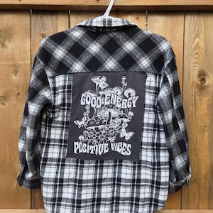 Può includere: Camicia di flanella a quadri bianchi e neri con una toppa sul retro. La toppa presenta un disegno di funghi e fiori con le parole "GOOD ENERGY" e "POSITIVE VIBES" in lettere bianche. La camicia è appesa a una gruccia bianca.