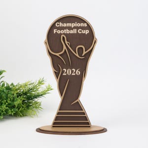 May include: A dark brown wooden trophy shaped like a stylized cup, featuring the engraved text "Champions Football Cup" and the year "2026" in a lighter wood tone. The trophy has a layered design with a circular base and a smooth finish.
