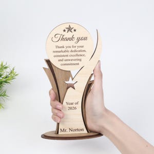 May include: A light-colored wooden trophy featuring a circular top with the text Thank you, followed by Thank you for your remarkable dedication, consistent excellence, and unwavering commitment. Below is a star cutout, Year of 2026, and Mr. Norton.