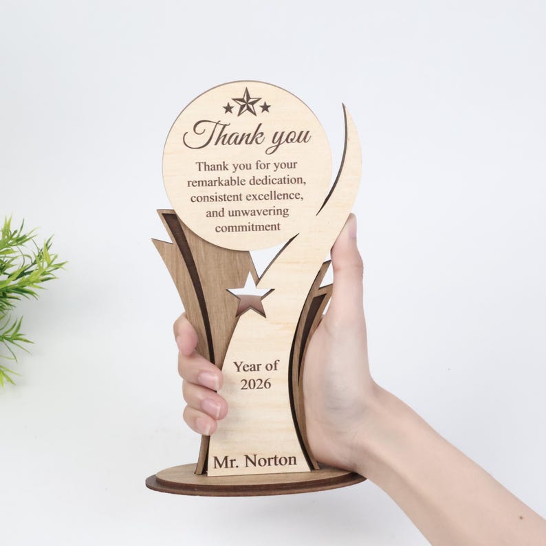 May include: A light-colored wooden trophy featuring a circular top with the text Thank you, followed by Thank you for your remarkable dedication, consistent excellence, and unwavering commitment. Below is a star cutout, Year of 2026, and Mr. Norton.