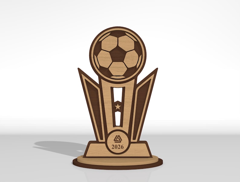 May include: A laser-cut wooden trophy featuring a soccer ball at the top, a central column with a star cutout, and a base displaying a triangular logo and the year 2026. The design uses contrasting light and dark wood tones with clean edges.