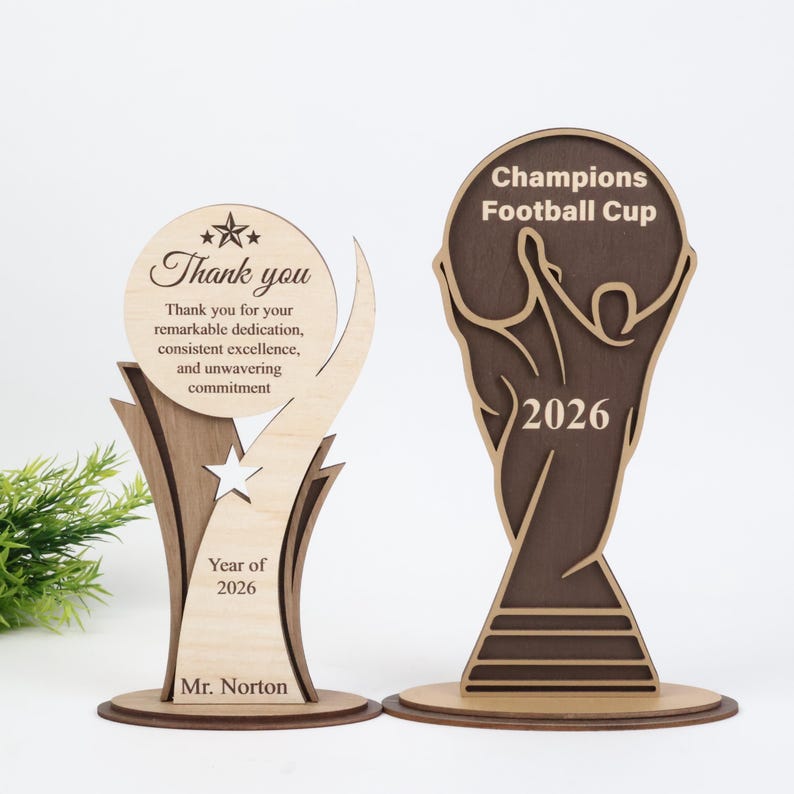 May include: Two wooden trophies with laser-cut designs. The left trophy is light wood with a circular top featuring a star and text: Thank you for your remarkable dedication, consistent excellence, and unwavering commitment. Year of 2026. Mr. Norton.
