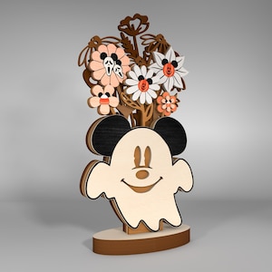 Puede incluir: Una decoración de Halloween de madera con un fantasma con orejas de Mickey Mouse y una cara sonriente. El fantasma está rodeado de un ramo de flores con pétalos de color naranja, rosa y blanco.