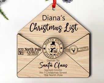 Könnte beinhalten: Ein hölzerner Weihnachtsanhänger in Form eines Umschlags mit einem Stempel des Nordpol-Postamts. Der Anhänger ist mit dem Namen "Diana's Christmas List" personalisiert und enthält die Adresse "Santa Claus, Santa's Grotto, No. 1 Christmas Street, The North Pole".