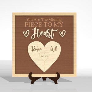 Puede incluir: Placa de madera con fondo marr&oacute;n y marco de madera clara. El texto "You Are The Missing Piece To My Heart" est&aacute; en la parte superior. Un rompecabezas en forma de coraz&oacute;n con los nombres "Robin" y "Will" est&aacute; en el centro. Tambi&eacute;n se incluye la fecha "25.12.2025".