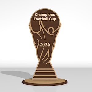 May include: A flat, laser-cut wooden trophy featuring a stylized figure with arms raised, holding a circular top. The text Champions Football Cup is engraved at the top, and the year 2026 is centered on the figure. The design uses layered dark and light wood tones.