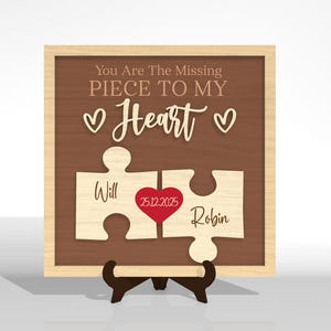 Puede incluir: Letrero de madera con dos piezas de rompecabezas. El letrero tiene el texto "You Are The Missing Piece To My Heart". Los nombres "Will" y "Robin" se muestran en las piezas del rompecabezas. Un coraz&oacute;n rojo con la fecha "25.12.2025" est&aacute; en el centro. El marco es de color marr&oacute;n claro.