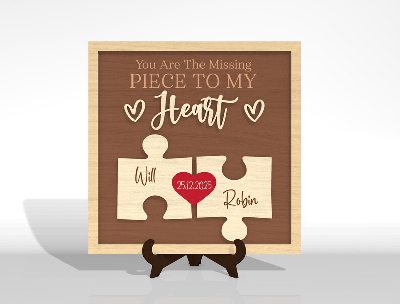 Puede incluir: Letrero de madera con dos piezas de rompecabezas. El letrero tiene el texto "You Are The Missing Piece To My Heart". Los nombres "Will" y "Robin" se muestran en las piezas del rompecabezas. Un coraz&oacute;n rojo con la fecha "25.12.2025" est&aacute; en el centro. El marco es de color marr&oacute;n claro.