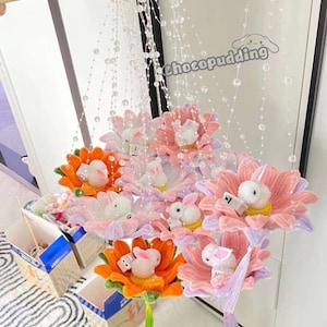 May include: A hanging mobile with multiple pastel-colored flower-shaped ornaments. Each flower has a small white bunny inside. The flowers are pink, purple, and orange. The mobile is decorated with clear beads and a blue and purple butterfly at the top.
