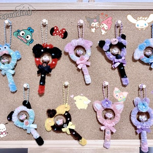 May include: A corkboard with 10 colorful pipe cleaner keychains with small glass beads inside. Each keychain has a different character or design, including a blue and green monster, a red and black Mickey Mouse, a pink and purple unicorn, and a blue and white bunny.