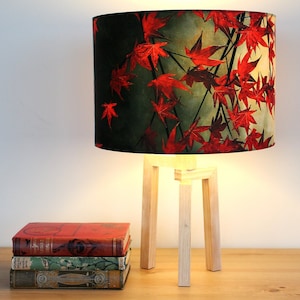 Peut inclure: Une lampe de table avec une base en bois blanc et un abat-jour cylindrique à imprimé floral rouge et vert. L'abat-jour présente un motif de feuilles d'érable rouges sur un fond vert foncé.
