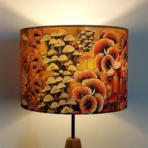 A drum lampshade on a table lamp base with warm light, featuring different kinds of mushrooms and fungi. The colours are earthy and autumnal with soft yellows, oranges, pinks, off white, black. A bit of a funky 70s feel, or maybe Miyazaki.