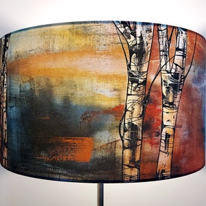 A large lampshade on a standard lamp. The lampshade design is painterly, with black and white birch trunks. The design shows visible brush strokes. It is multicoloured with earthy red/brown to the right, and blues, yellow and copper to the left.