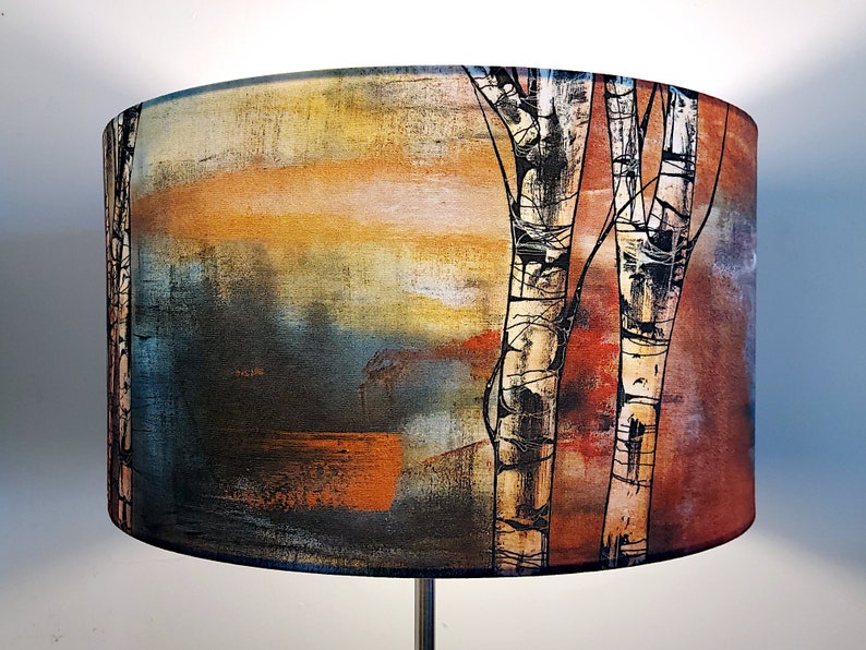 A large lampshade on a standard lamp. The lampshade design is painterly, with black and white birch trunks. The design shows visible brush strokes. It is multicoloured with earthy red/brown to the right, and blues, yellow and copper to the left.