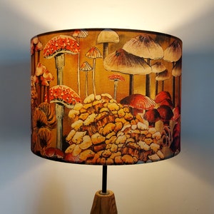 A drum lampshade on a table lamp base with warm light, featuring different kinds of mushrooms and fungi. The colours are earthy and autumnal with soft yellows, oranges, pinks, off white, black. A bit of a funky 70s feel, or maybe Miyazaki.