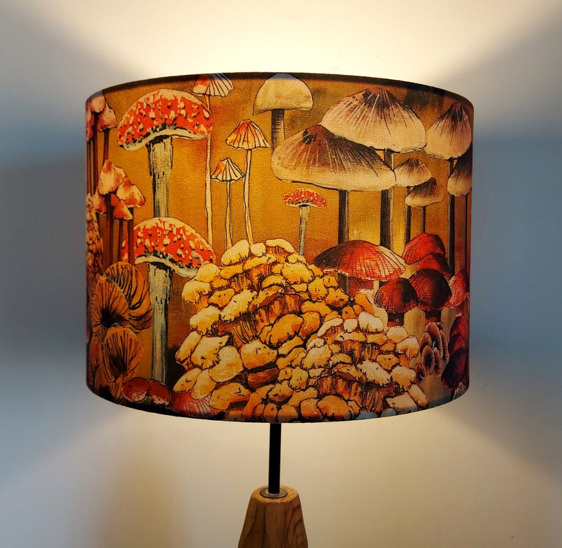A drum lampshade on a table lamp base with warm light, featuring different kinds of mushrooms and fungi. The colours are earthy and autumnal with soft yellows, oranges, pinks, off white, black. A bit of a funky 70s feel, or maybe Miyazaki.