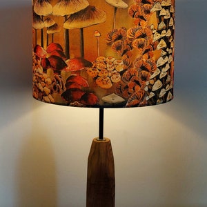A drum lampshade on a wooden lamp base with warm light, featuring different kinds of mushrooms and fungi. The colours are earthy and autumnal with soft yellows, oranges, pinks, off white, black. A bit of a funky 70s feel, or maybe Miyazaki.