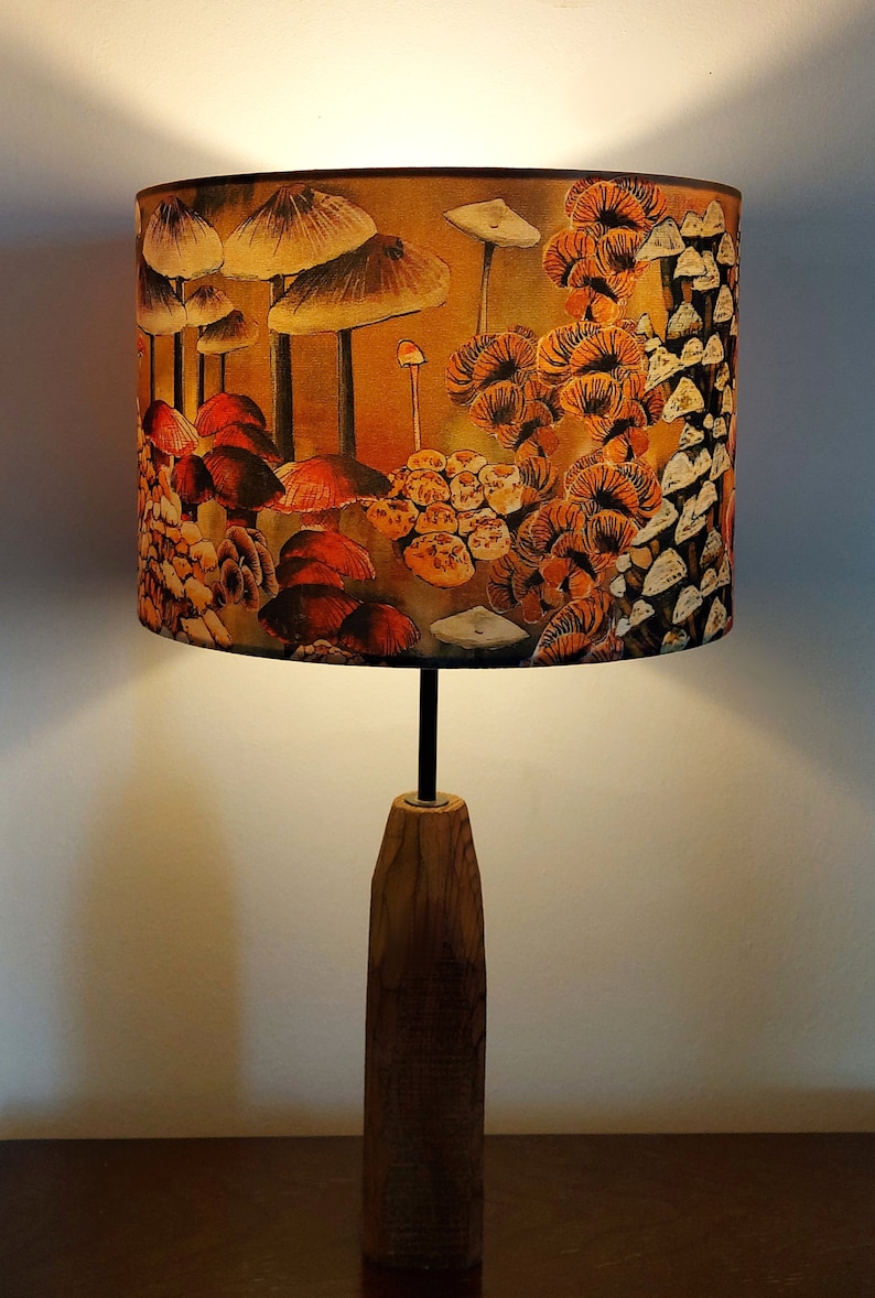 A drum lampshade on a wooden lamp base with warm light, featuring different kinds of mushrooms and fungi. The colours are earthy and autumnal with soft yellows, oranges, pinks, off white, black. A bit of a funky 70s feel, or maybe Miyazaki.