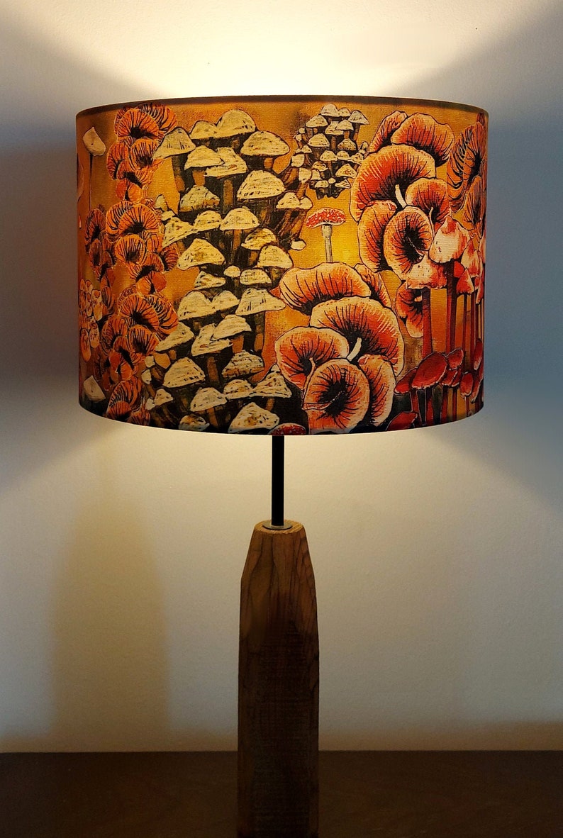 A drum lampshade on a table lamp base with warm light, featuring different kinds of mushrooms and fungi. The colours are earthy and autumnal with soft yellows, oranges, pinks, off white, black. A bit of a funky 70s feel, or maybe Miyazaki.