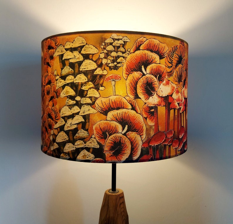 A drum lampshade on a table lamp base with warm light, featuring different kinds of mushrooms and fungi. The colours are earthy and autumnal with soft yellows, oranges, pinks, off white, black. A bit of a funky 70s feel, or maybe Miyazaki.