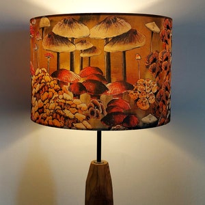 A drum lampshade on a table lamp base with warm light, featuring different kinds of mushrooms and fungi. The colours are earthy and autumnal with soft reds, yellows, oranges, pinks, off white, black. A bit of a funky 70s feel, or maybe Miyazaki.