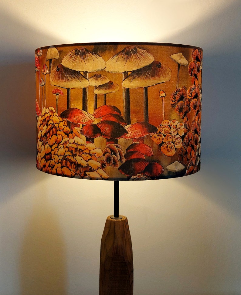 A drum lampshade on a table lamp base with warm light, featuring different kinds of mushrooms and fungi. The colours are earthy and autumnal with soft reds, yellows, oranges, pinks, off white, black. A bit of a funky 70s feel, or maybe Miyazaki.