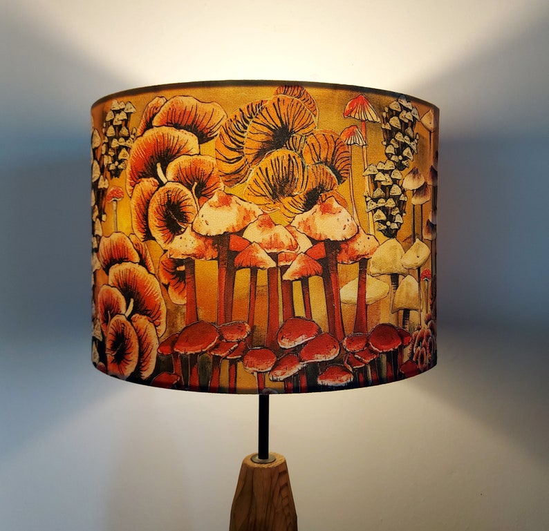 A drum lampshade on a table lamp base with warm light, featuring different kinds of mushrooms and fungi. The colours are earthy and autumnal with reds, yellows, oranges, pinks, off white, black. A bit of a funky 70s feel, or maybe Miyazaki.