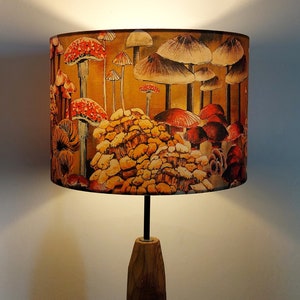 A drum lampshade on a table lamp base with warm light, featuring different kinds of mushrooms and fungi. The colours are earthy and autumnal with reds, yellows, oranges, pinks, off white, black. A bit of a funky 70s feel, or maybe Miyazaki.
