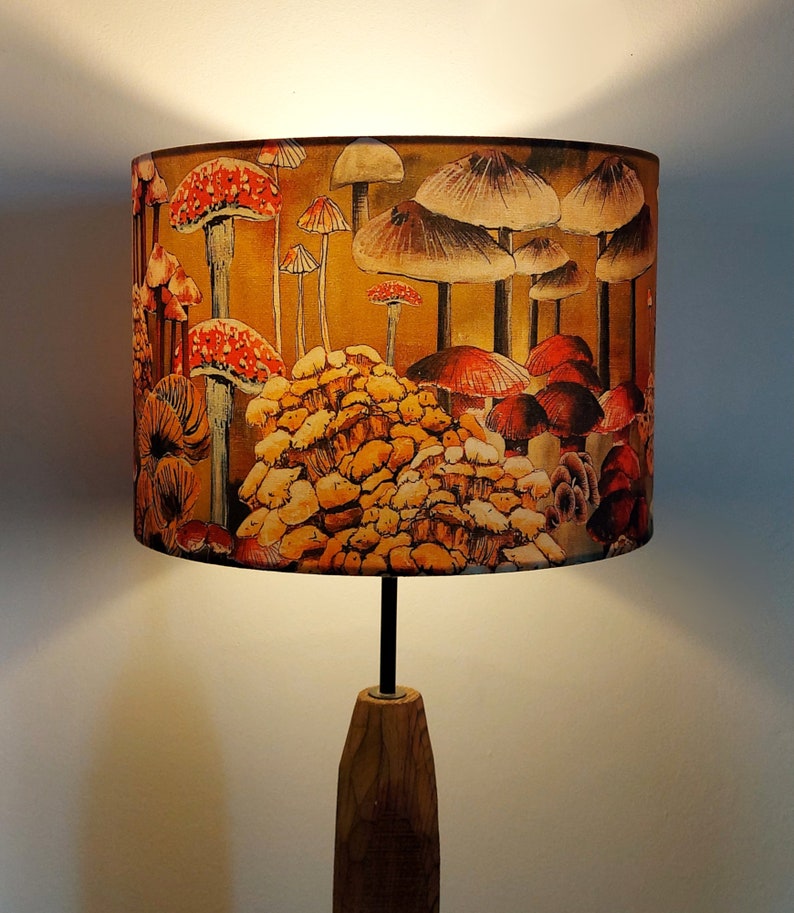 A drum lampshade on a table lamp base with warm light, featuring different kinds of mushrooms and fungi. The colours are earthy and autumnal with reds, yellows, oranges, pinks, off white, black. A bit of a funky 70s feel, or maybe Miyazaki.