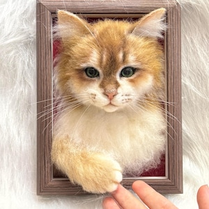 May include: A framed felted cat portrait. The cat has golden-brown fur with white patches, green eyes, and a gentle expression. The frame is a warm brown, and the background is a soft, textured white.