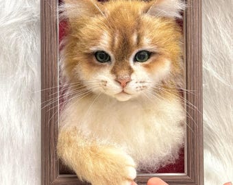 Op de afbeelding: Een ingelijst vilten kattenportret. De kat heeft goudbruine vacht met witte accenten, groene ogen en een lieve uitdrukking. De lijst is warmbruin en de achtergrond is zacht, getextureerd wit.