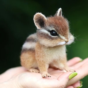 Puede incluir: Una figura de ardilla listada en miniatura, realista, con pelaje marrón y beige, posada sobre una palma abierta. La ardilla tiene ojos oscuros, hocico blanco y bigotes. El fondo es verde borroso.