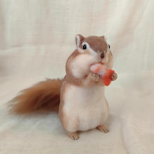 May include: A handcrafted felt squirrel figurine in shades of brown, tan, and white, holding a small, orange object. The squirrel has a fluffy tail and is posed upright, giving it a lifelike appearance. The background is a soft, off-white fabric.