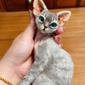 May include: A handmade, gray cat doll with large, expressive teal eyes and oversized ears. The cat has a slender build and is held in a person's hand. The doll's fur appears soft and textured, with a detailed face and whiskers.