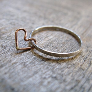 May include: A silver ring with a small copper heart charm hanging from it. The ring is on a wooden surface.