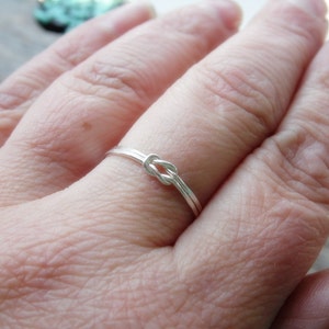 May include: A silver knot ring on a finger. The ring is simple and delicate, with a smooth finish.