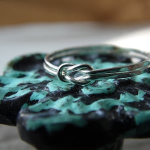 May include: A silver ring with a knot design, resting on a teal and black surface.