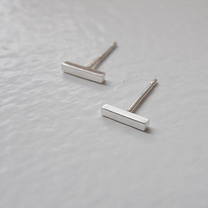 May include: A pair of silver bar stud earrings. The earrings are simple and minimalist, with a rectangular bar shape.
