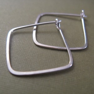 Peut inclure: Une paire de boucles d'oreilles créoles carrées en argent. Les boucles d'oreilles sont en métal fin et ont un design simple et moderne.