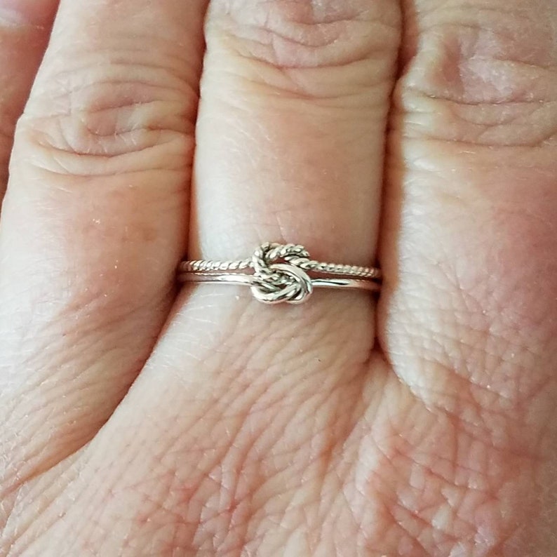 May include: A silver ring with a knot design. The ring is worn on a finger.