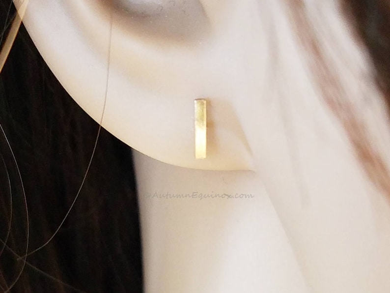 May include: A close-up of a gold bar earring. The earring is a small, rectangular shape and is positioned on the ear. The background is blurred, with a focus on the earring.