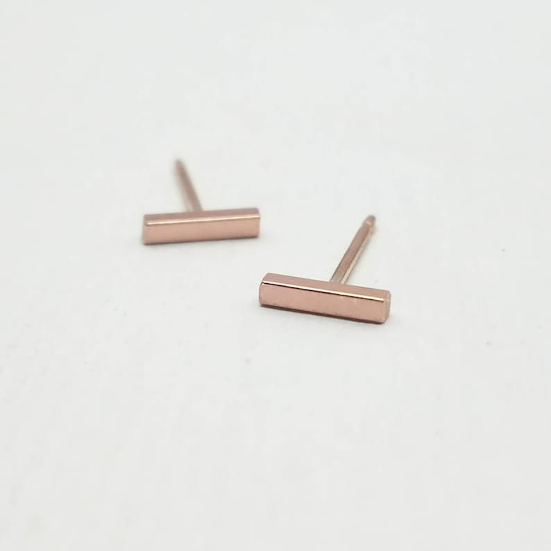 May include: Two rose gold bar stud earrings. Each earring features a small, rectangular bar shape with a post for insertion. The earrings are displayed on a white surface, showcasing their simple, modern design. These earrings are a minimalist jewelry accessory.