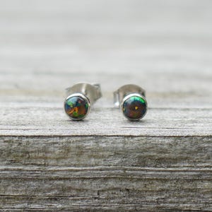 May include: A pair of stud earrings featuring round, dark-toned opals with vibrant, multi-coloured flecks of green, orange, and red. The stones are set in polished silver-coloured metal settings with standard post backs for secure attachment.