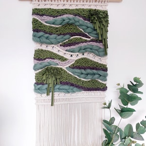 Könnte beinhalten: Ein Wandbehang aus gewebtem Garn in Grün-, Lila- und Weißtönen. Der Behang hat unten einen Fransenbesatz und dekorative Quasten in Grün.