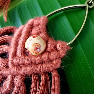 May include: Close-up of a handmade macramé earring. The earring features a rust-coloured woven design with a decorative bead and a brass-coloured metal hoop. The earring is set against a green leaf background.