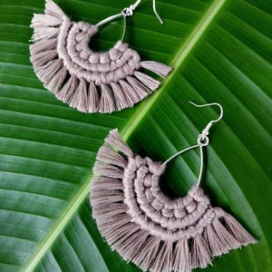 May include: A pair of light brown macrame earrings with a fan-shaped design and silver-coloured hooks. The earrings are displayed on a large, green leaf, highlighting the intricate knotting and fringe details. The earrings are handmade.