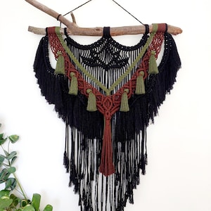 Puede incluir: Un tapiz de macramé para colgar en la pared con una combinación de colores negro, marrón y verde oliva. El diseño incluye intrincados nudos, borlas y una rama de madera natural para colgar. Esta pieza artesanal añade un toque bohemio.