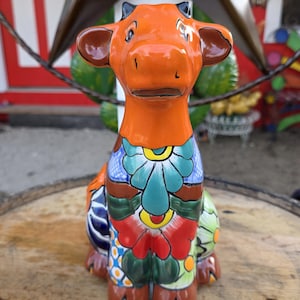 May include: A ceramic cow figurine with an orange body and brown ears. The cow is adorned with colorful floral patterns in blue, green, red, and yellow. The figurine is sitting on a wooden surface.